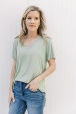 Model wearing jeans with a light sage short sleeve top with a v-neck and rayon/spandex blend.