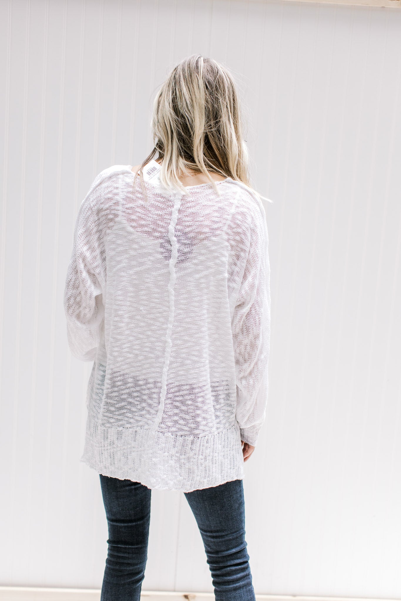 Back view of Model wearing a semi-sheer cream loose knit sweater with long sleeves and exposed hem. 