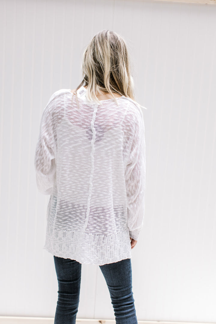 Back view of Model wearing a semi-sheer cream loose knit sweater with long sleeves and exposed hem. 