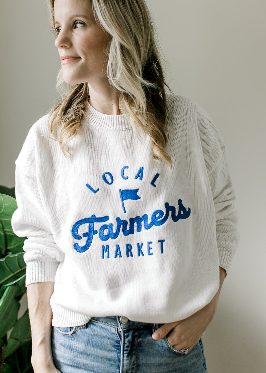 Model wearing a white sweater with bright blue Farmers Market logo, long sleeves and a round neck.