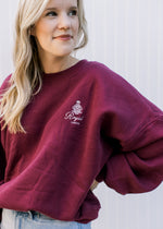 Model wearing a maroon sweatshirt with a cream royal London emblem, long sleeves and fleece lining. 