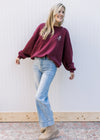 Model wearing jeans, booties and a maroon sweatshirt with a cream emblem and long sleeves. 