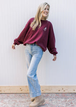 Model wearing jeans, booties and a maroon sweatshirt with a cream emblem and long sleeves. 