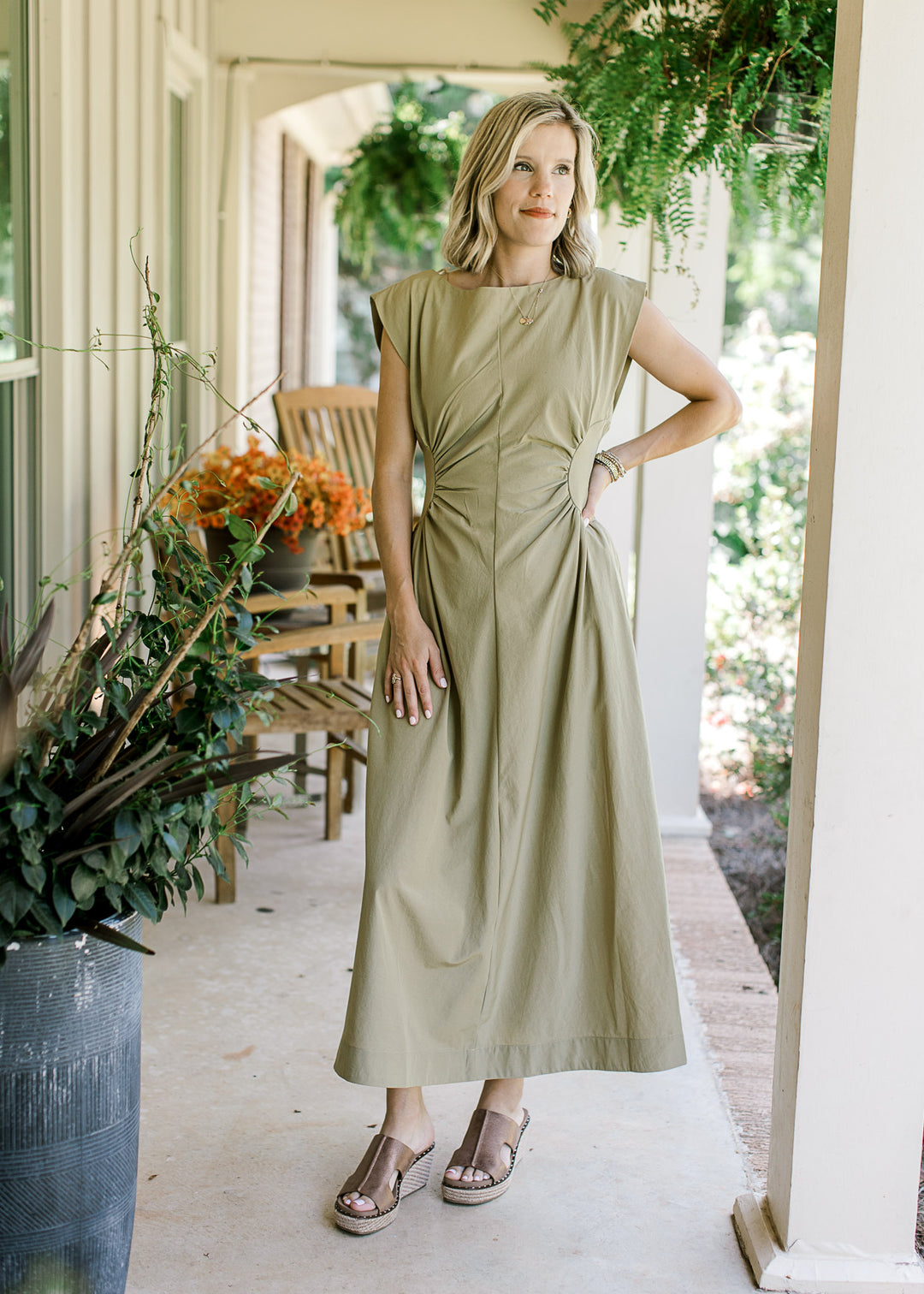 Model wearing a mossy tan sleeveless maxi with hidden zipper and ruched side panels at the waist. 