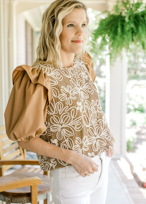 Model wearing a camel colored top with white floral and short puff sleeves with shoulder ruffle. 