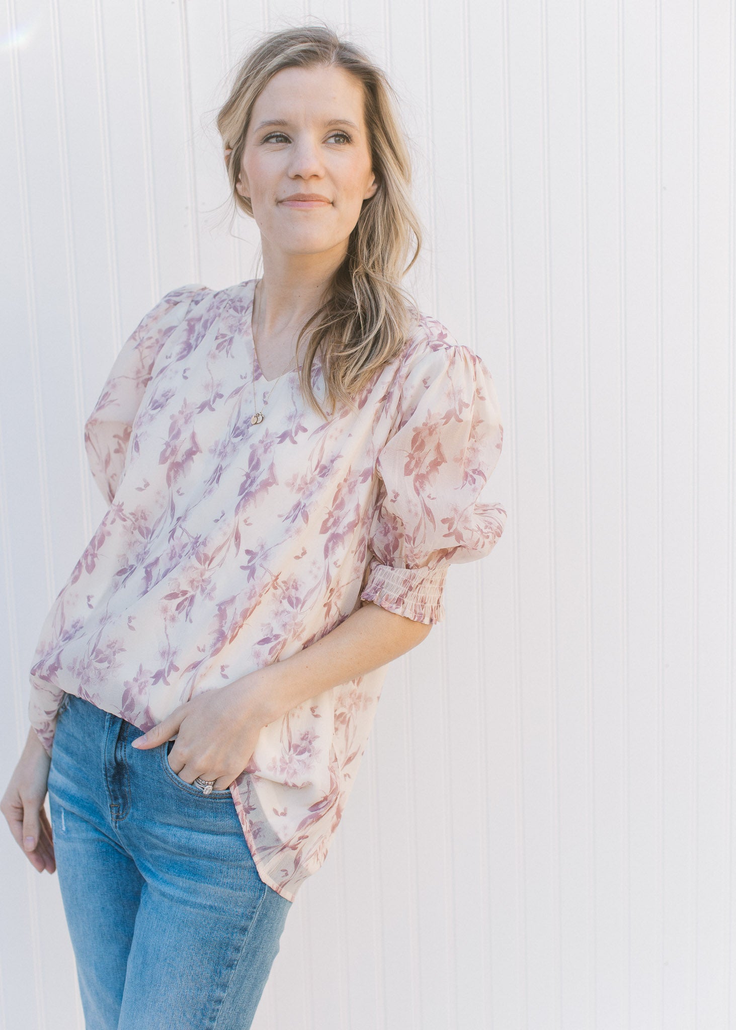 Model wearing jeans with a cream top with a mauve floral pattern, short sleeves and a v-neck.