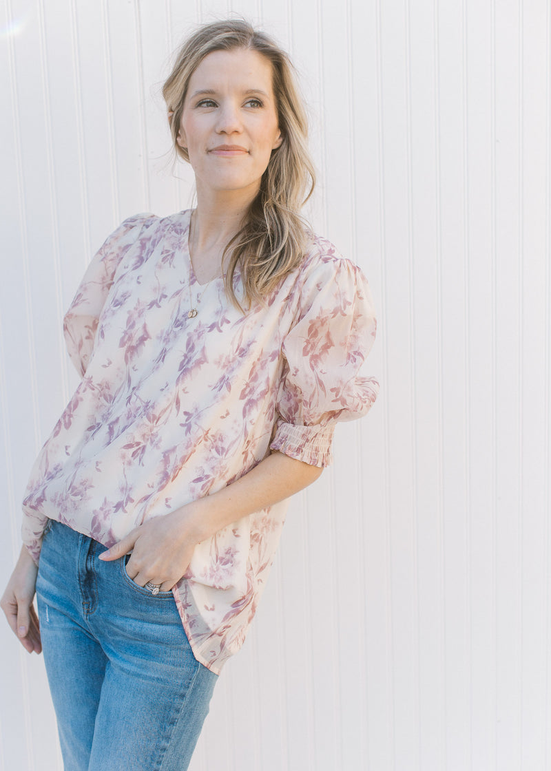 Model wearing jeans with a cream top with a mauve floral pattern, short sleeves and a v-neck.