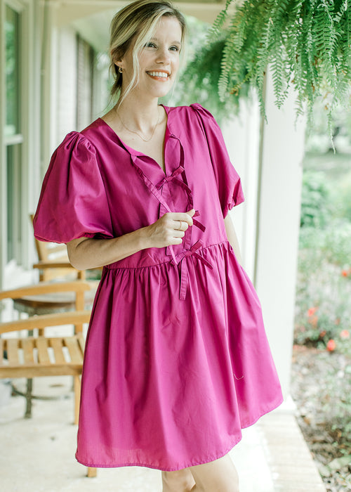 Model wearing a magenta above the knee dress with short puff sleeves and bow tie front. 