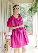 Model wearing a magenta dress with short puff sleeves, pockets and bow tie front closure. 