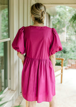 Back view of Model wearing a magenta dress with short puff sleeves and bow tie front closure. 