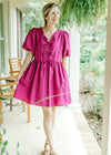 Model wearing sandals with a magenta dress with short puff sleeves and bow tie front closure. 