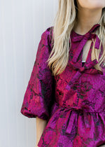 Model wearing a bubble 3/4 sleeve top with magenta flowers, peplum fit and bows down front.