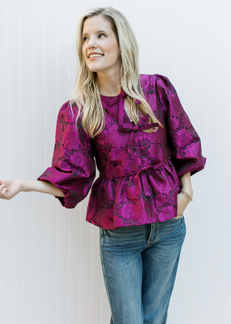 Model wearing jeans and a bubble 3/4 sleeve top with magenta flowers and bows down front.