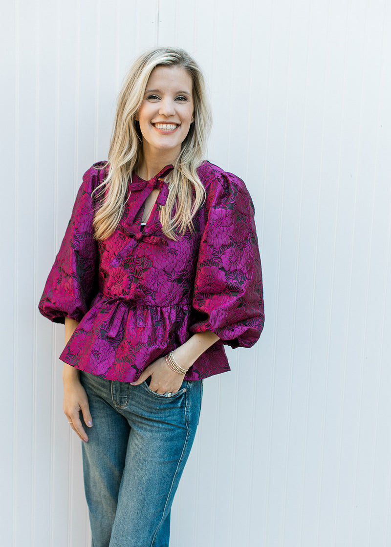 Model wearing a magenta floral v-neck top with bows down the front and 3/4 bubble sleeves.