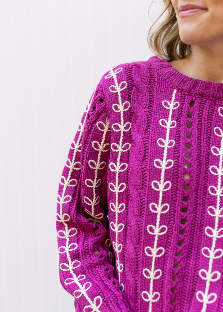 Close up of openwork knit and cream embroidery on a magenta sweater with a round neck. 
