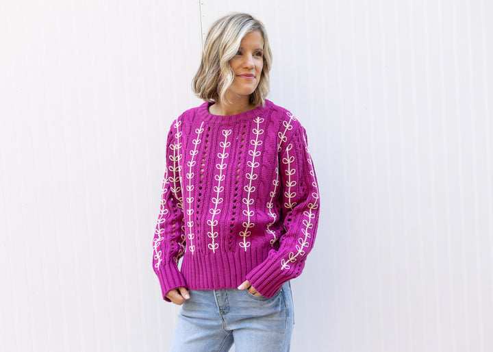 Model wearing an openwork knit magenta sweater with cream embroidered design and long sleeves.