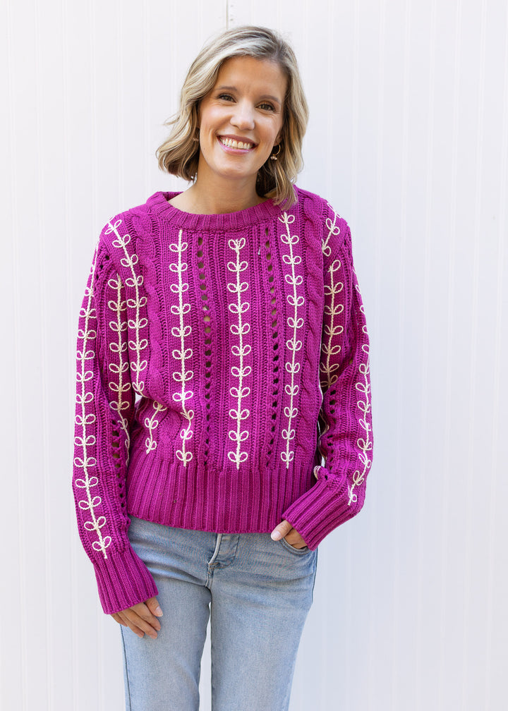 Model wearing jeans and an openwork knit magenta sweater with cream embroidery and long sleeves.