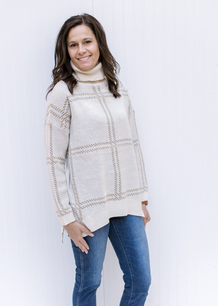 Model wearing jeans with a camel on cream plaid sweater with a turtleneck and long sleeves. 