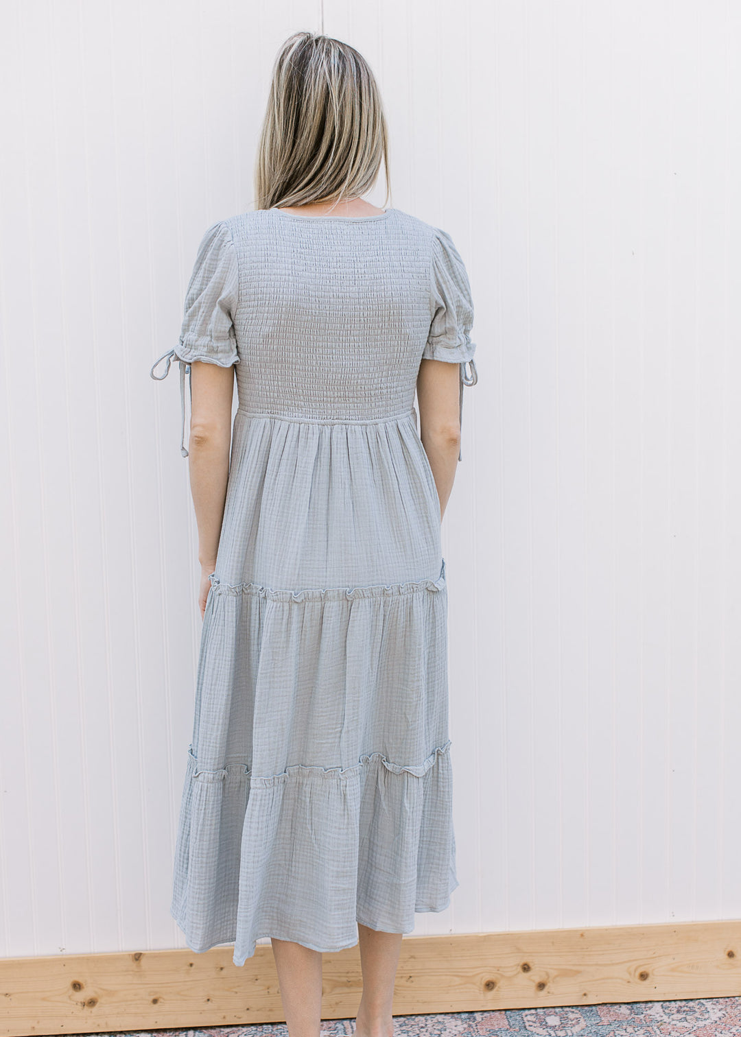 Back view of Model wearing a pale blue midi with smocked bodice and short sleeves with tie detail. 