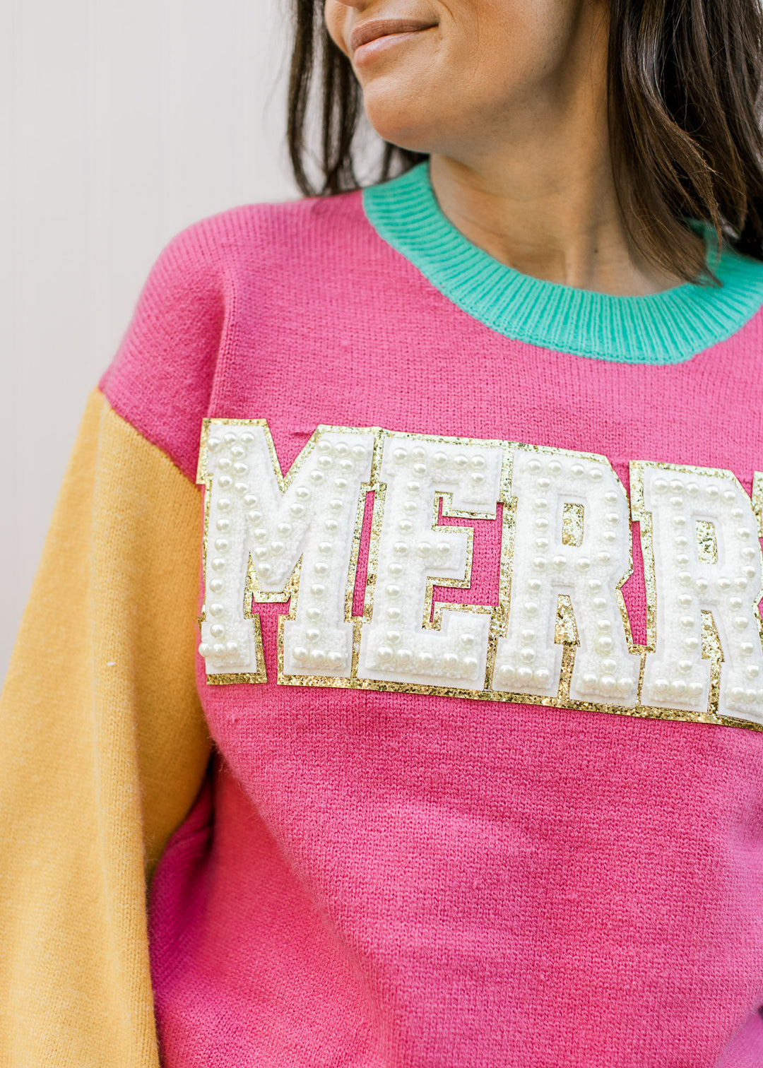 Close up of the word MERRY in cream outlined in gold on a pink long sleeve sweater. 