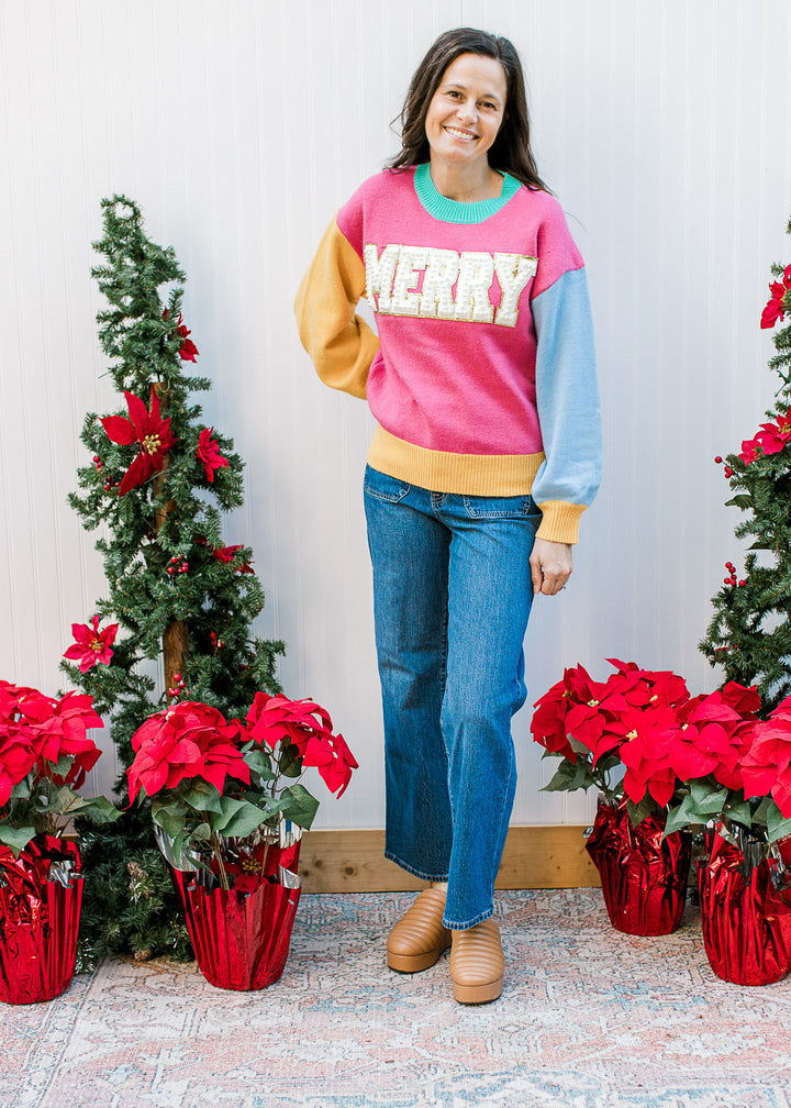 Model wearing jeans and a bright colorblock  “MERRY” sweater with a round neck and long sleeves. 