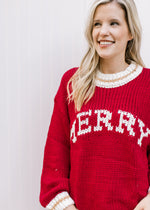 Model wearing a red long sleeve sweater with “MERRY” across the front in cream font. 