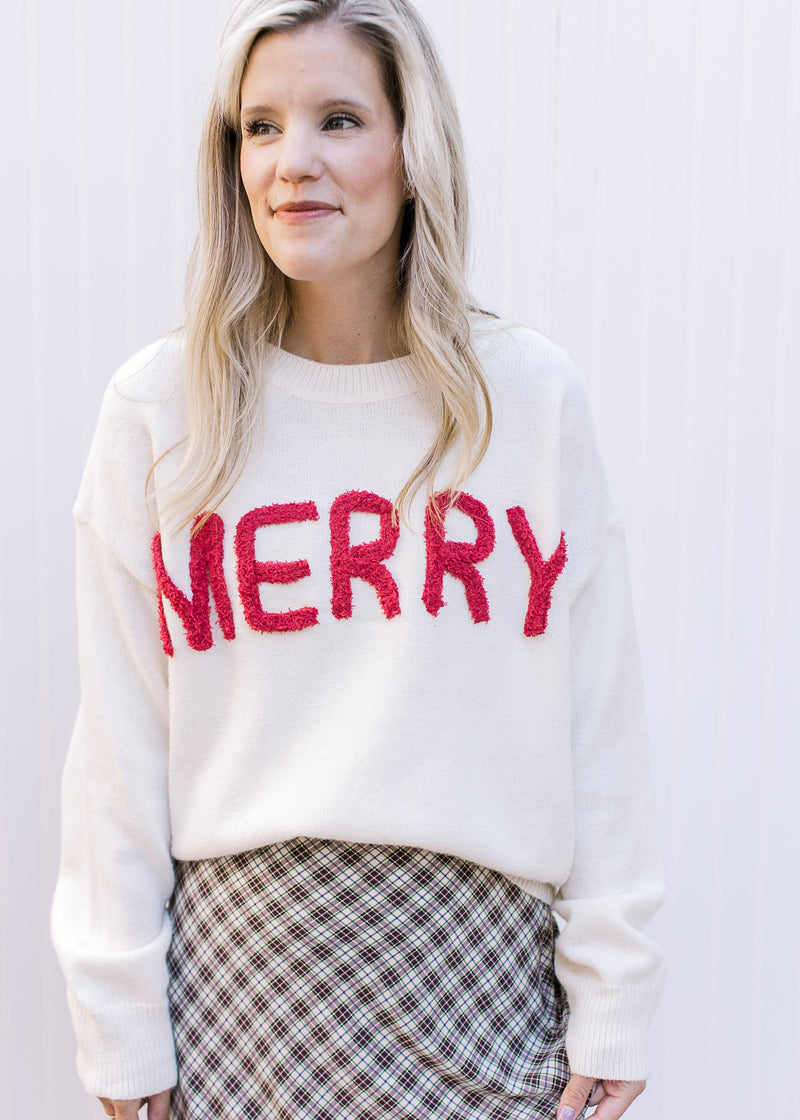 Model wearing a cream sweater with MERRY across the front in red yarn and long sleeves.