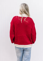 Back view of Model wearing a red long sleeve sweater with cream and gold at neck, hem and cuff. 