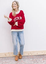 Model wearing jeans, clogs and a red sweater with “MERRY” across the front in cream font. 