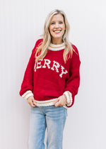 Model wearing a red sweater with “MERRY” across the front in cream font and long sleeves. 