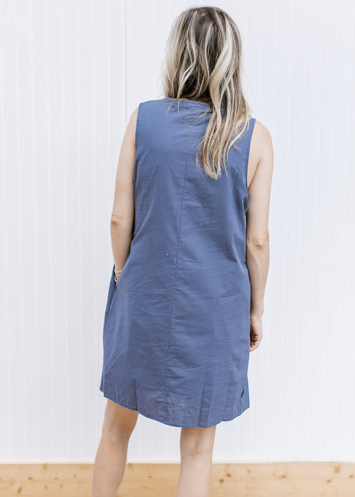 Back view of Model wearing a sleeveless navy dress with a pleated bodice, pockets and tie v-neck.
