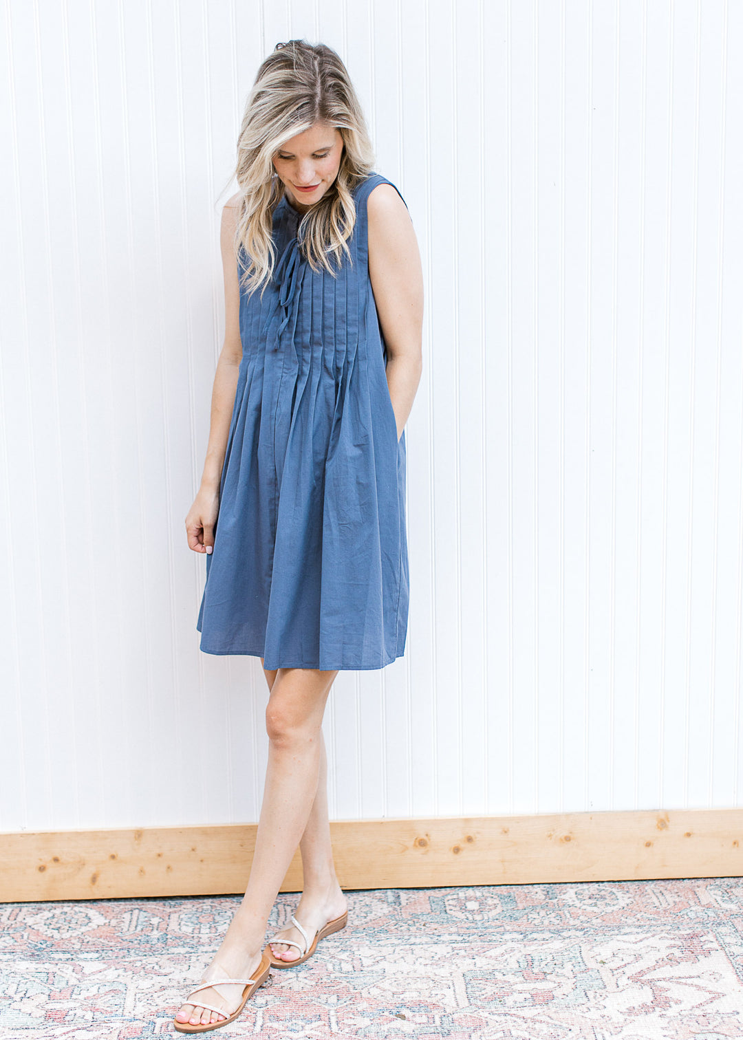Model wearing sandals and a sleeveless navy dress with a pleated bodice and double tie v-neck.
