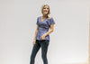 Model wearing jeans with a blue/violet top with gathered peplum and short flutter sleeves. 