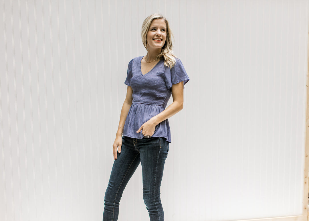 Model wearing jeans with a blue/violet top with gathered peplum and short flutter sleeves. 