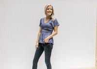 Model wearing jeans with a blue/violet top with gathered peplum and short flutter sleeves. 