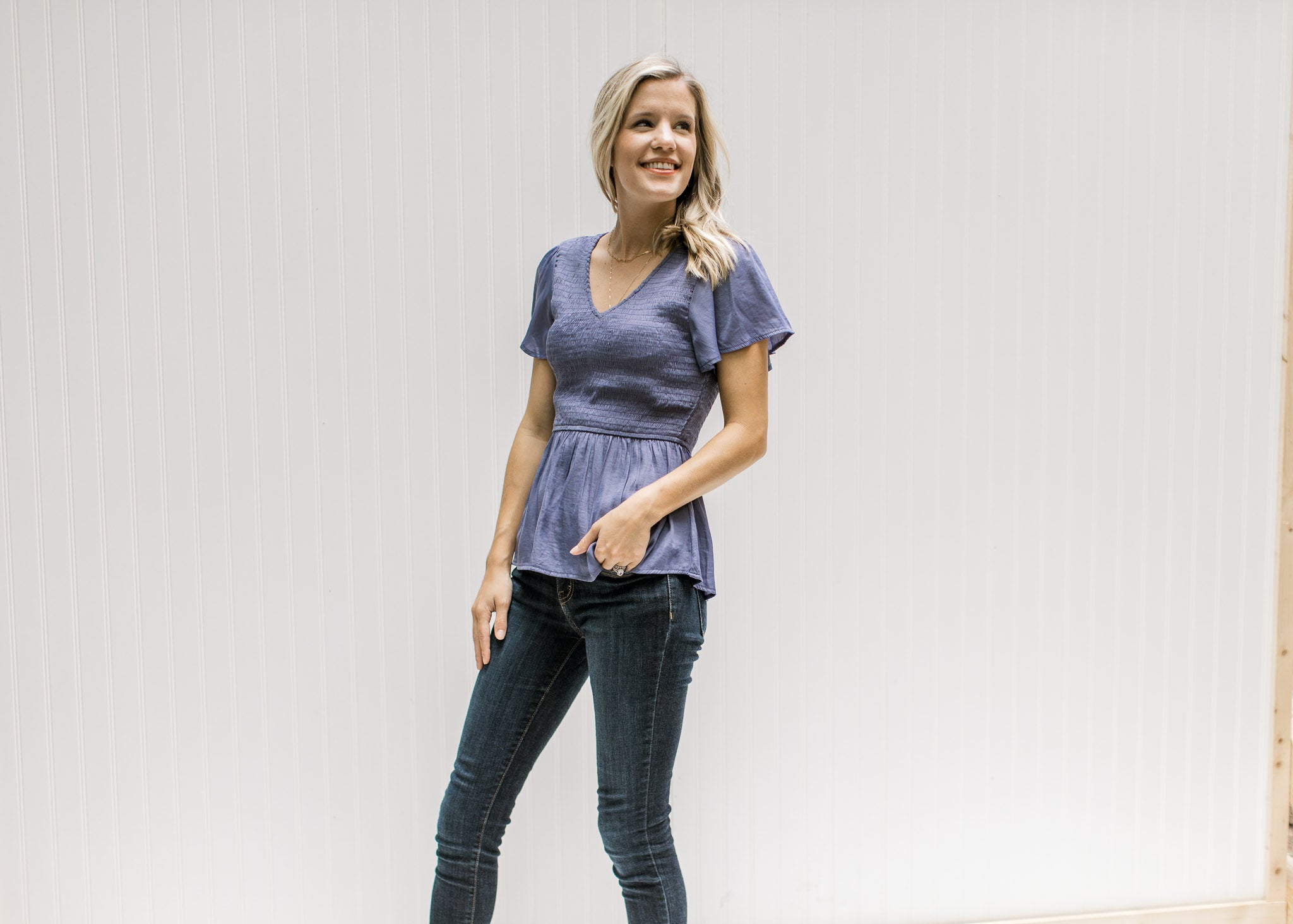 Model wearing jeans with a blue/violet top with gathered peplum and short flutter sleeves. 