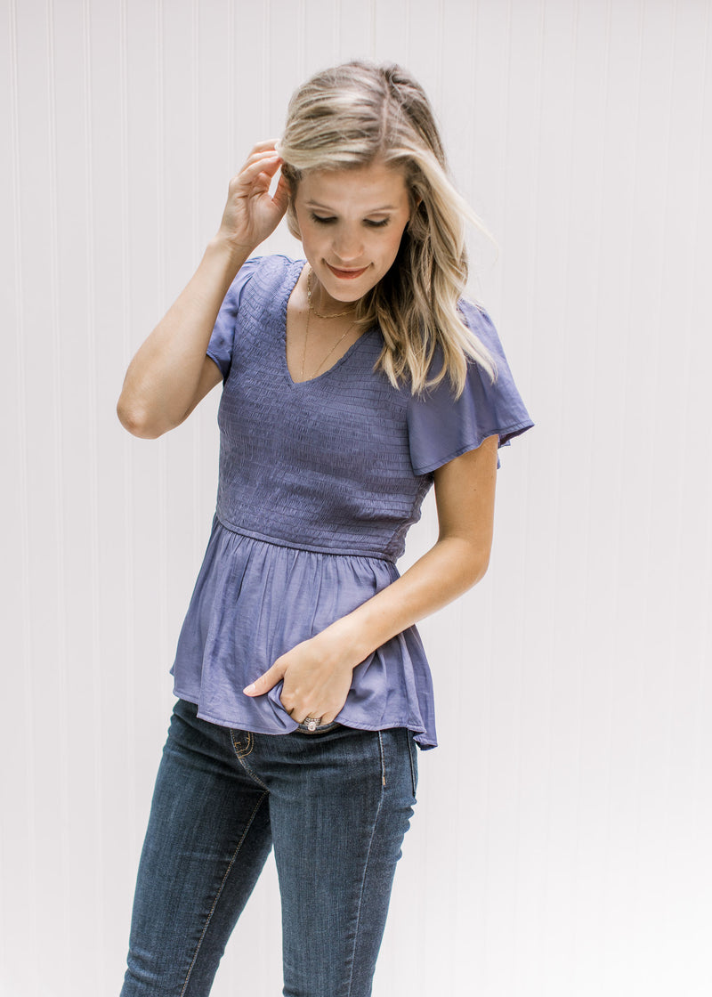 Model wearing a blue/violet colored v-neck top with a smocked bodice and short flutter sleeves. 