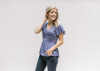 Model wearing a blue/violet top with gathered peplum, smocked bodice and short flutter sleeves. 