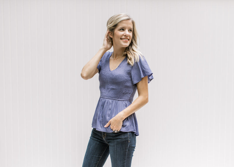 Model wearing a blue/violet top with gathered peplum, smocked bodice and short flutter sleeves. 
