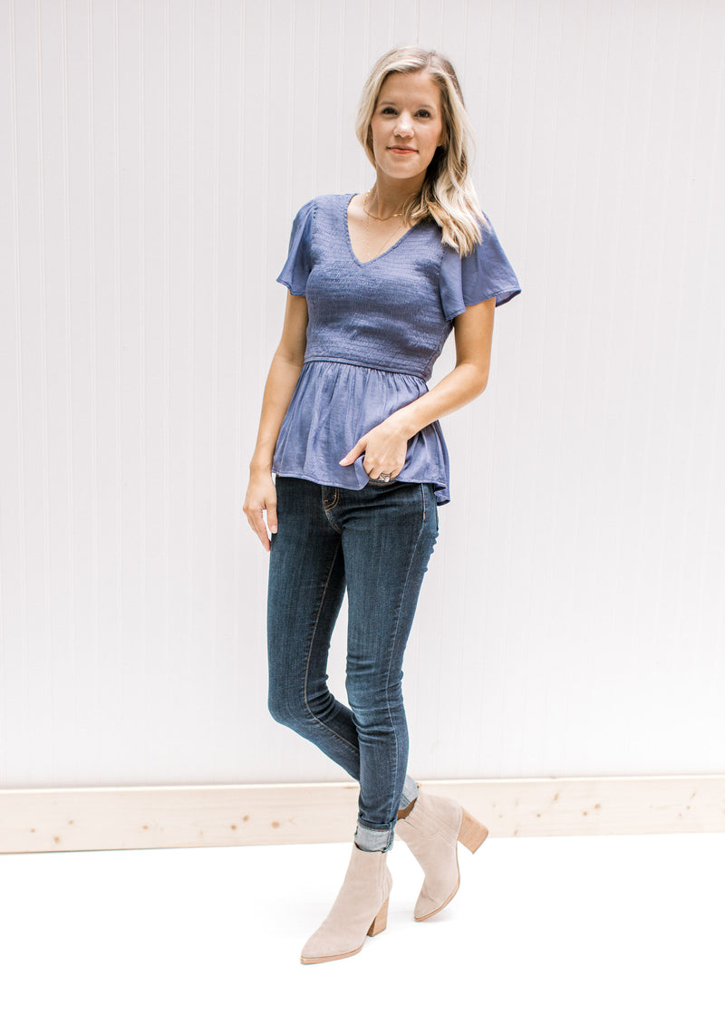 Model wearing jeans and booties with a blue/violet top with a smocked bodice and short sleeves.