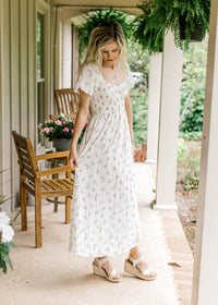 Model wearing wedges and a cream maxi with lavender floral, a smocked bodice and short sleeves.