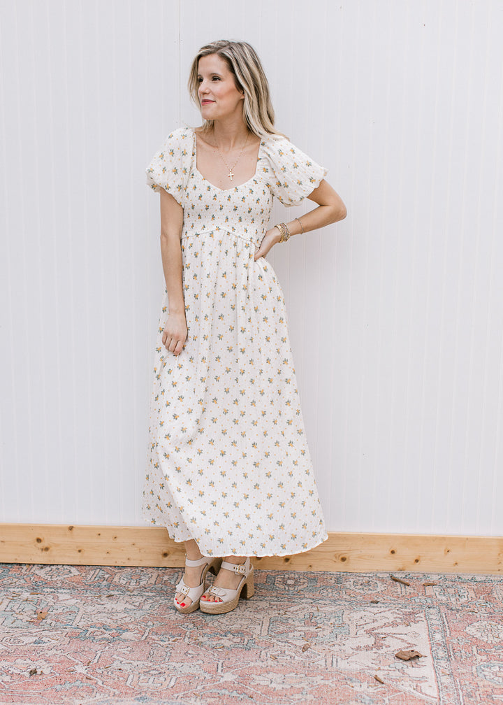 Model wearing a necklace, heels and a cream maxi with yellow floral and smocked bodice. 