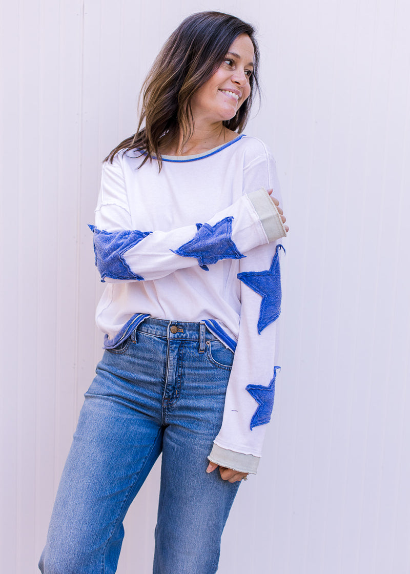 Model wearing a cream top with blue patch stars along the long sleeve and exposed hem. 