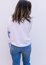 Back view of Model wearing a cream top with blue patch stars along the long sleeve and exposed hem. 