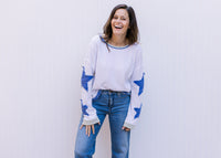 Model wearing jeans and a cream top with blue patch stars along the long sleeve and exposed hem. 