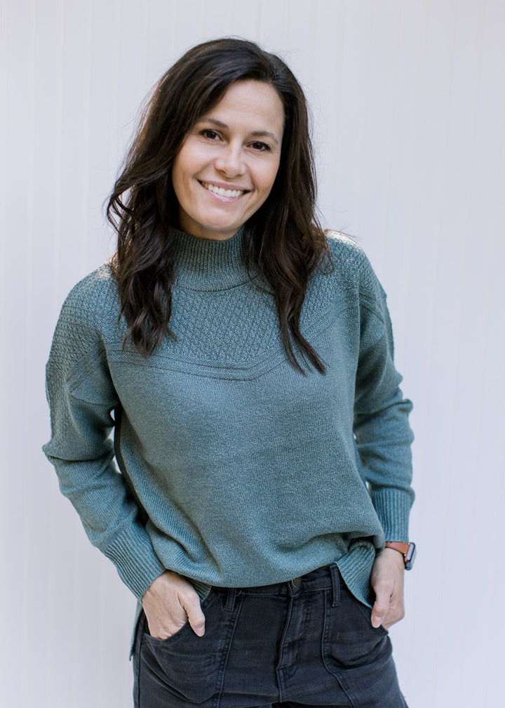 Model wearing a mottled sage green sweater with a mock neck, long sleeves and ribbed neck and cuff.