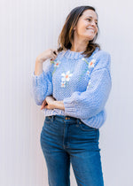 Model wearing jeans and a soft blue knit sweater with white flowers, 3/4 sleeves and a round neck.