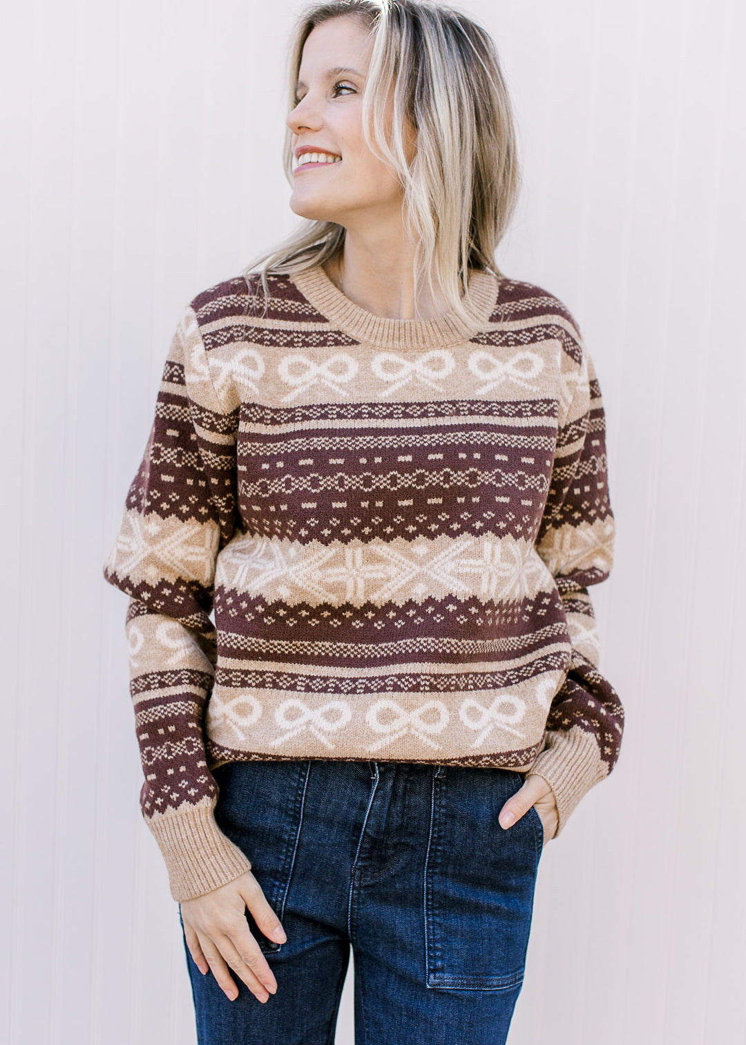 Model wearing a tan and brown sweater with cream bow accents, round neck and long sleeves. 