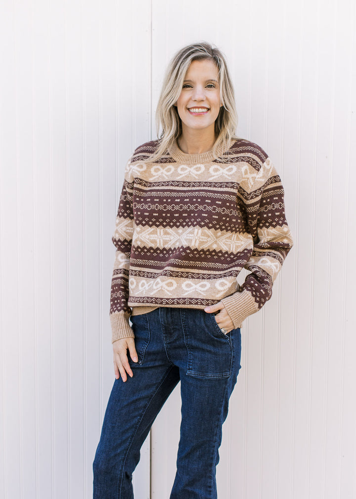 Model wearing jeans and a tan and brown patterned sweater with cream bow accents and long sleeves. 