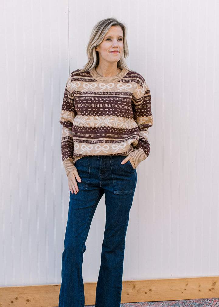 Model wearing a cozy tan and brown patterned sweater with cream bow accents and long sleeves. 